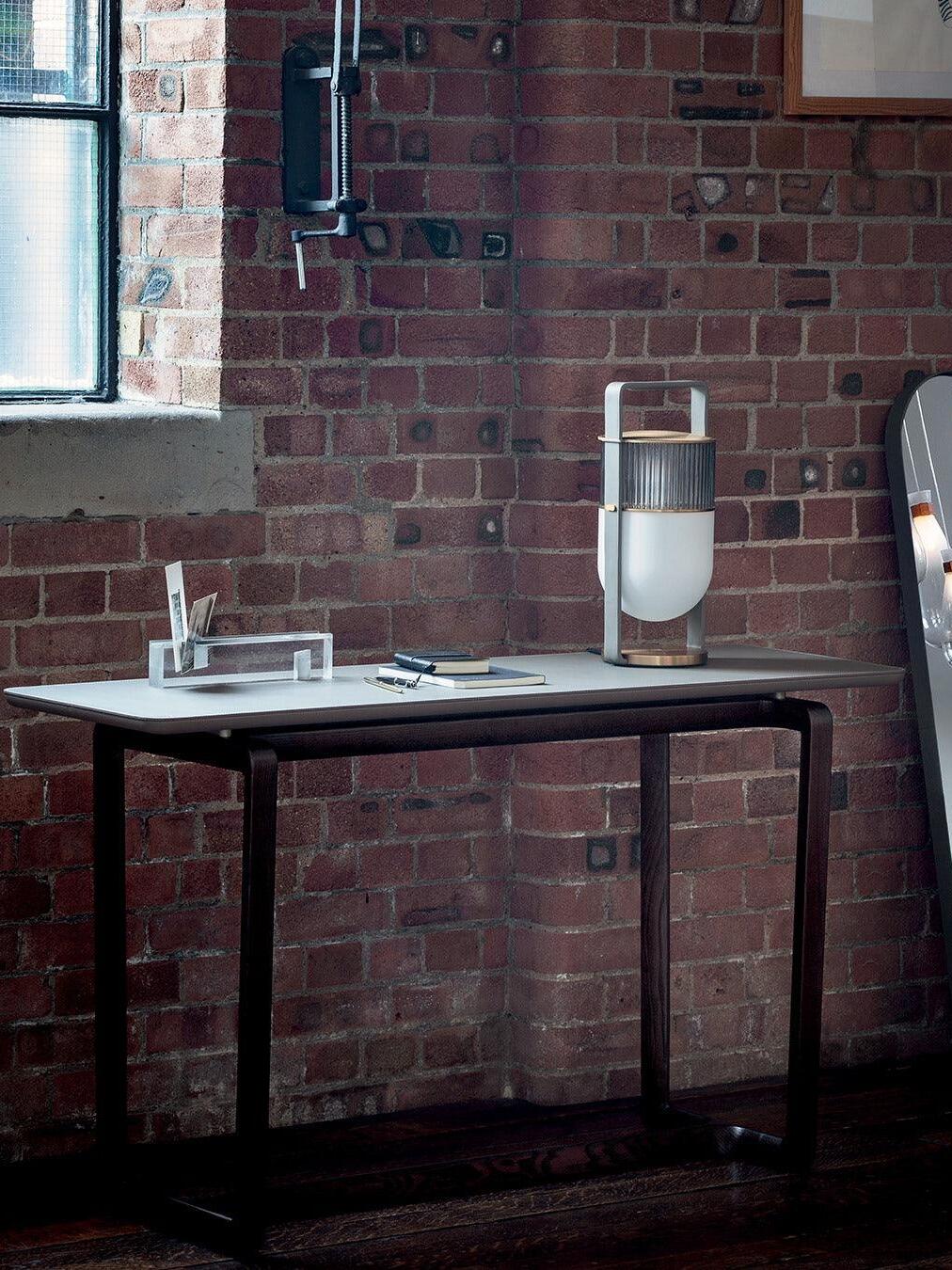 Xi lamp on a desk by an exposed brick wall and window, with books and a drinking glass
