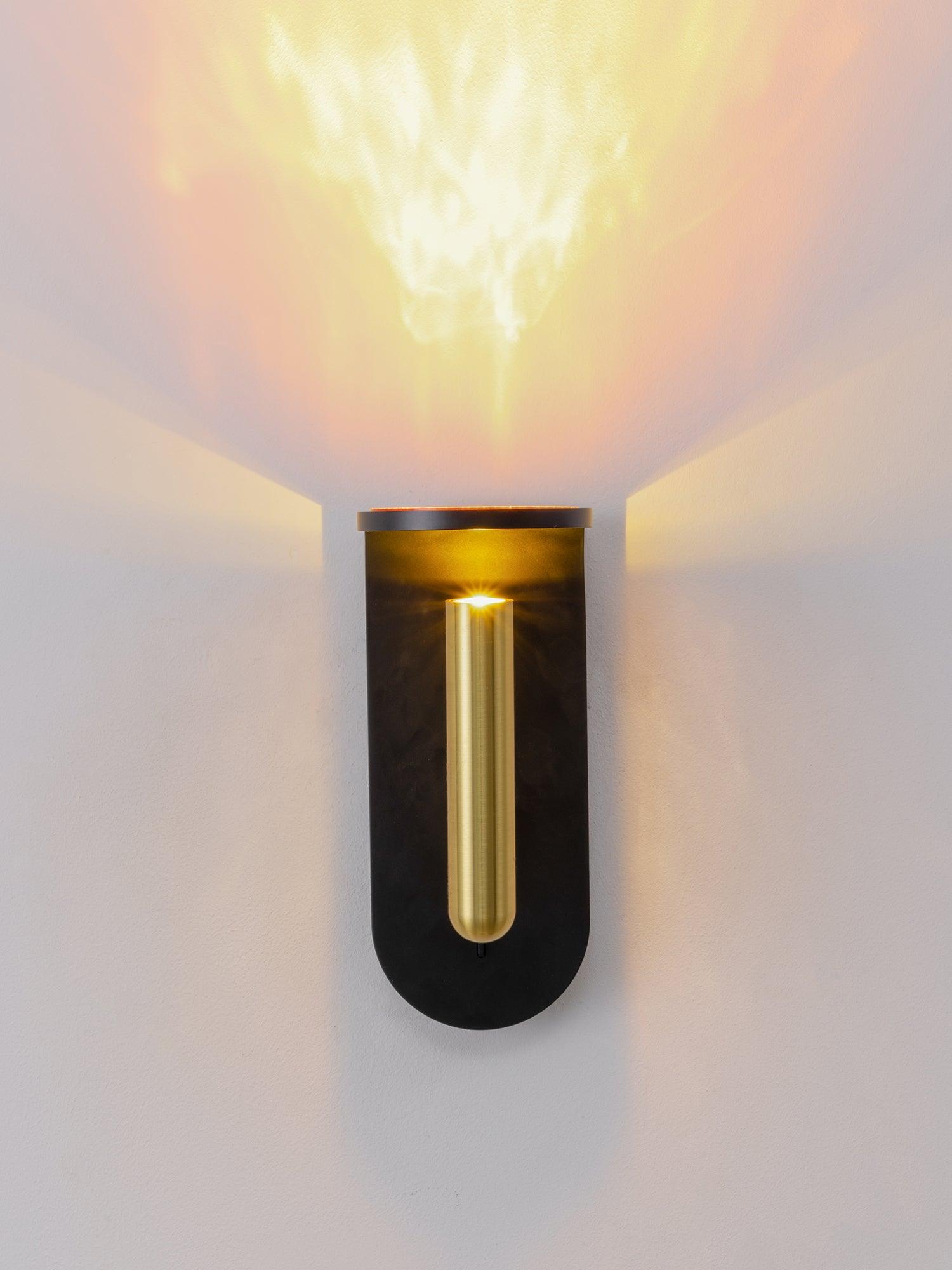 Front view of a black Nights wall light with a vertical gold-tone head and bright pattern above it