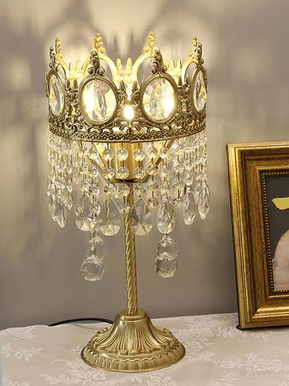 Illuminated crown-shaped crystal table lamp with long lower drops beside a framed picture.