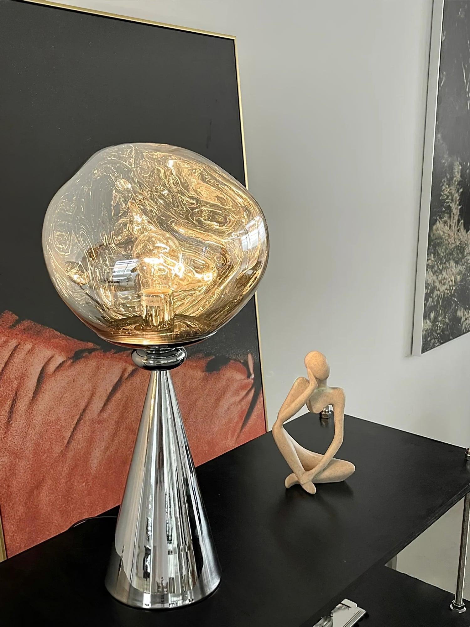 Glowing chrome globe table lamp on a black cabinet beside a seated wooden figure and red artwork