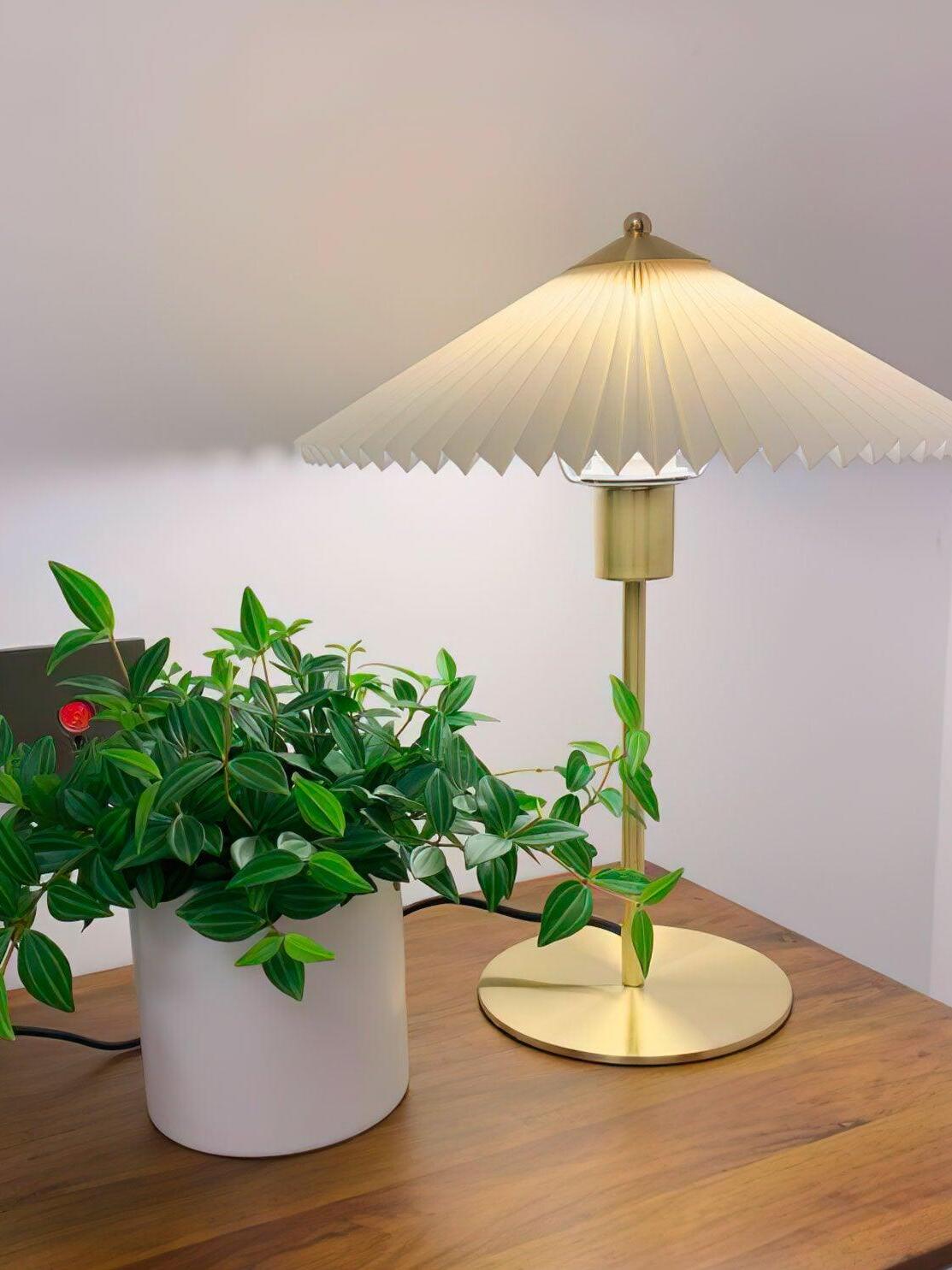 Gold-tone Perla table lamp glowing beside a leafy plant on a wooden surface