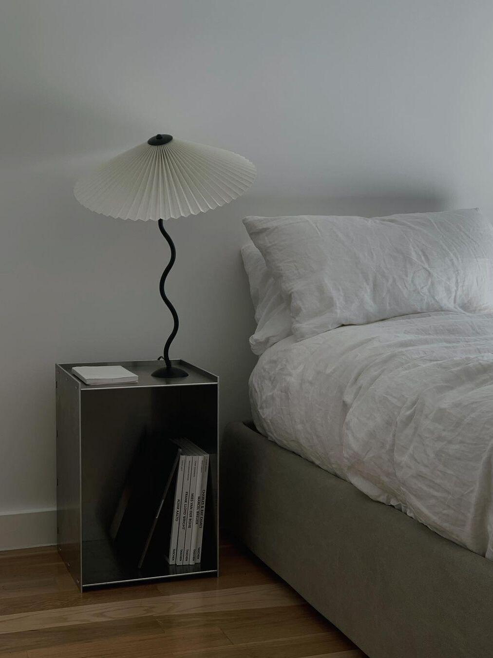 White pleated-shade lamp on an open metal bedside cabinet beside a white bed and books