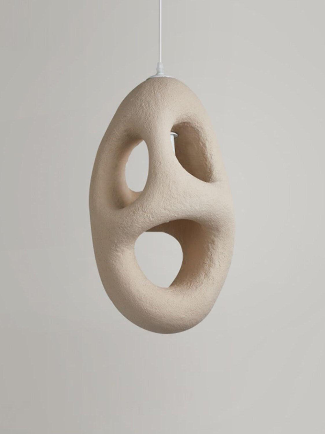 Unlit elongated cream-looking textured pendant with three large through-openings and White cord against Gray