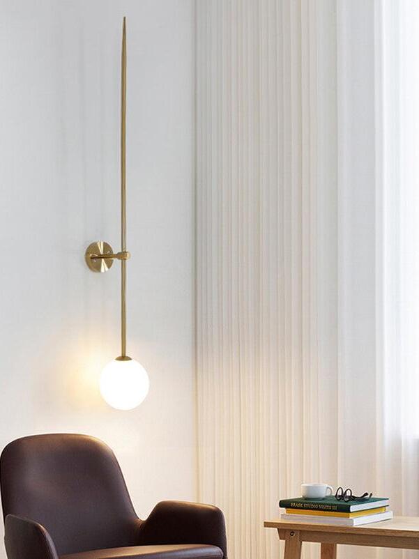 Lit gold-tone rod wall light beside a brown armchair, books and a cup near white curtains