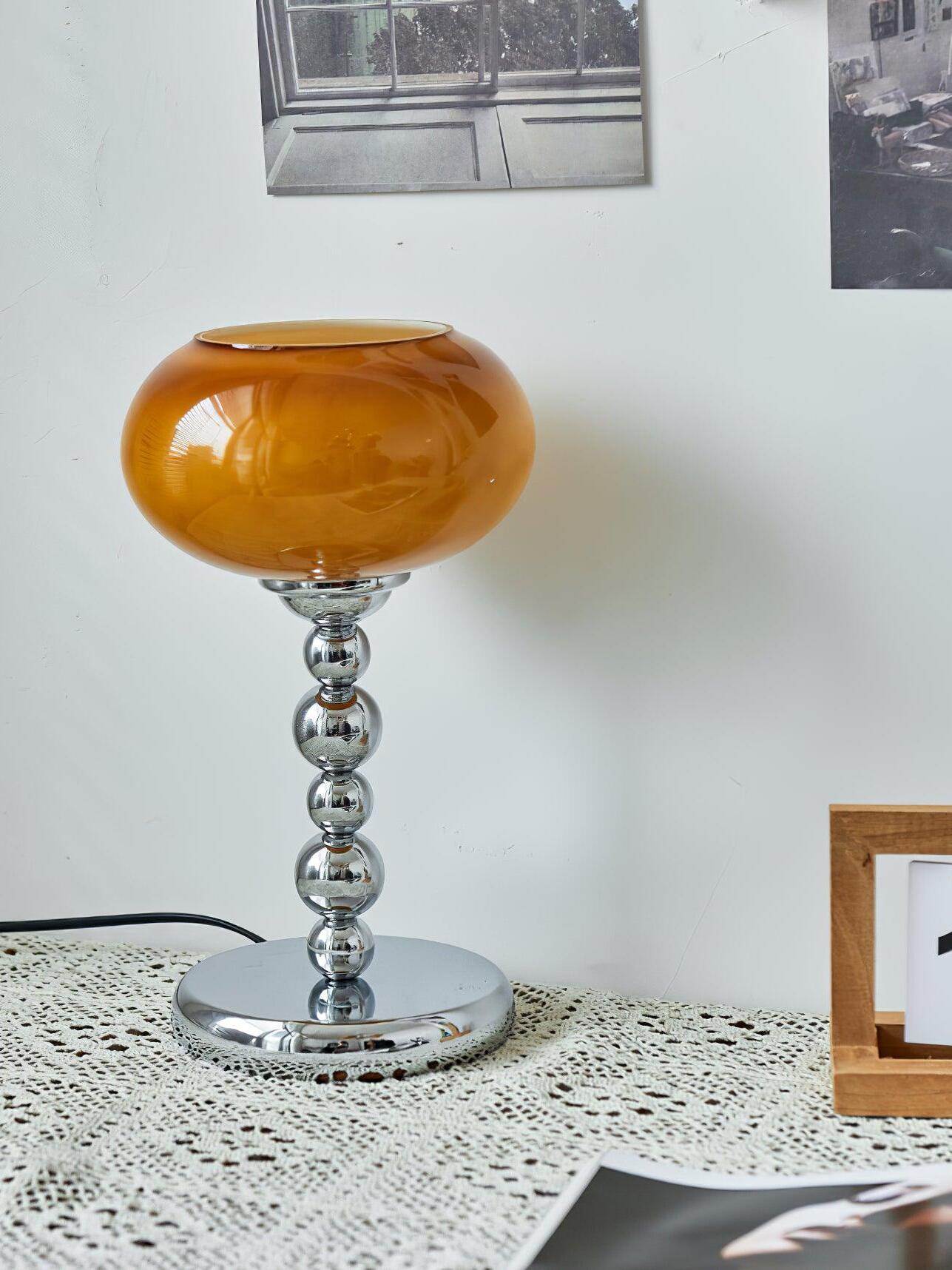 Front view of the unlit dark-orange glass table lamp on lace cloth, with a rounded chrome stem and framed art behind