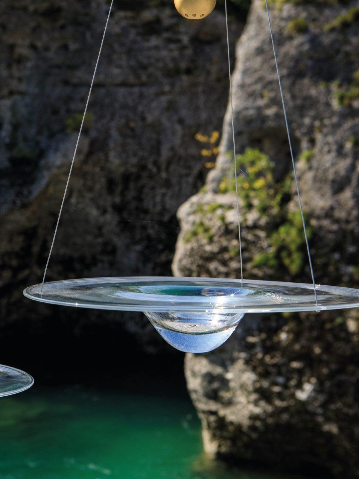 Clear glass disc pendant photographed against rock and water