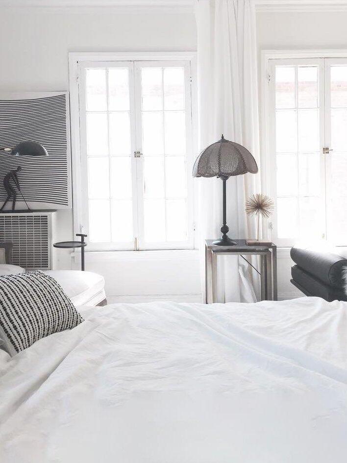 White bedroom with a partly cropped black dome light at left and a separate dark scalloped table lamp near the windows