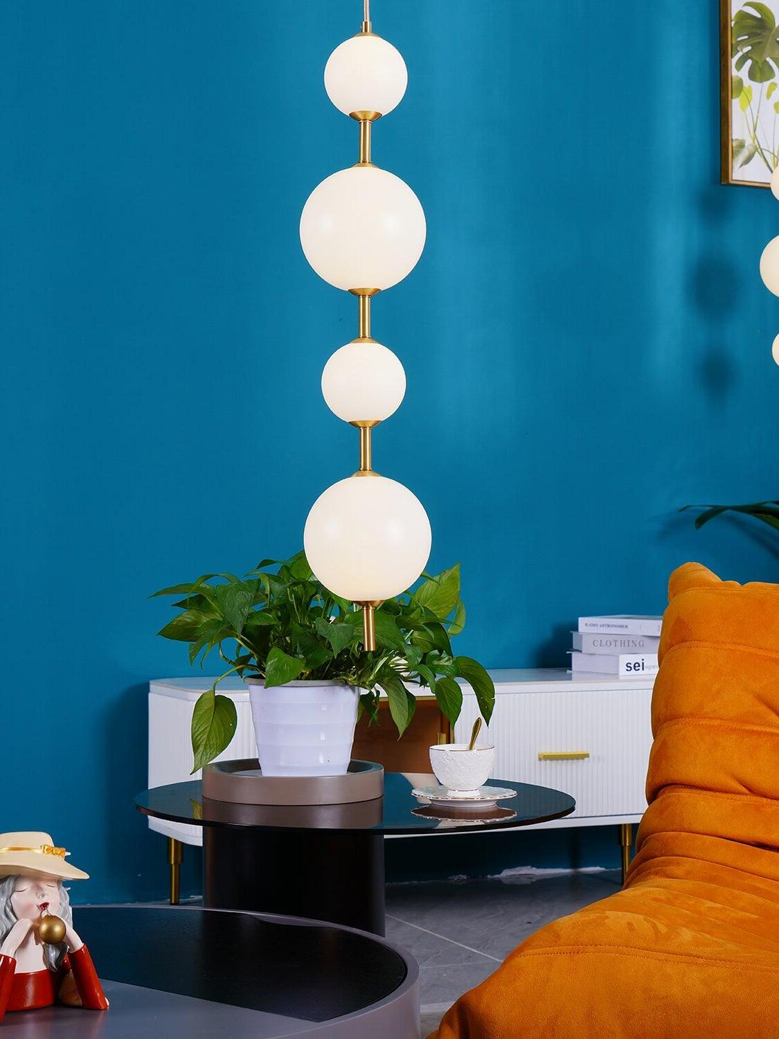 Four glowing white globes with brass-tone links against a teal wall near a cabinet and orange chair