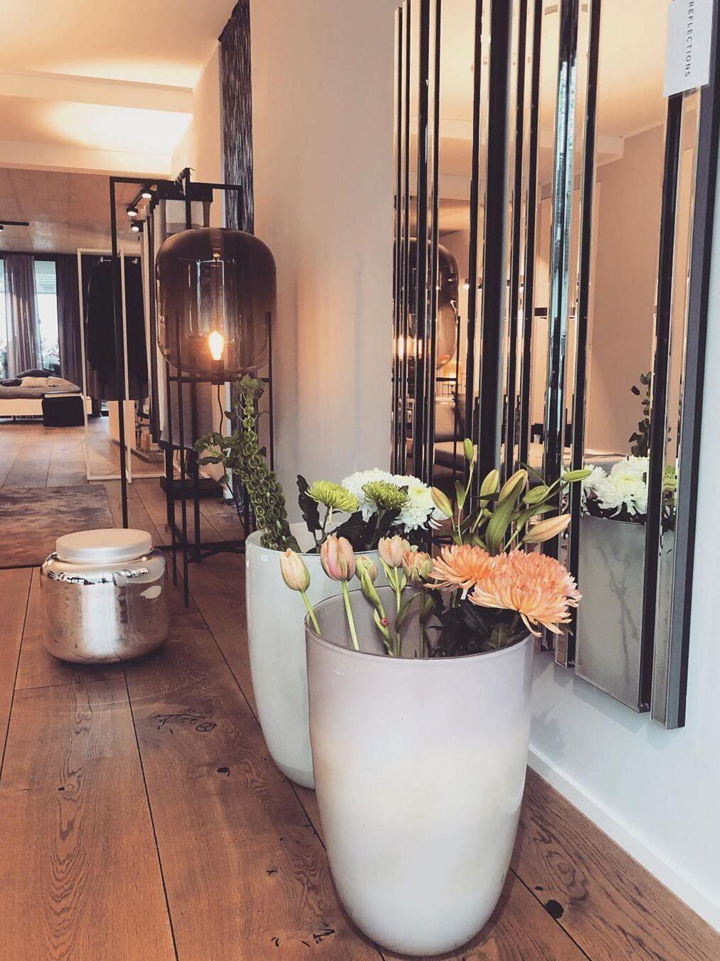Illuminated glass floor lamp beside wall mirrors and a vase of flowers.
