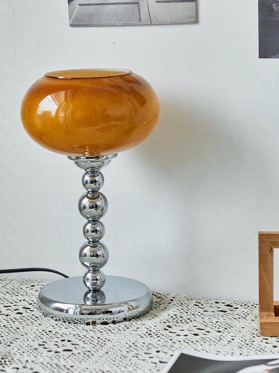 Dark-orange oval glass table lamp on lace cloth beside framed art, with a rounded chrome stem, circular base and black cord