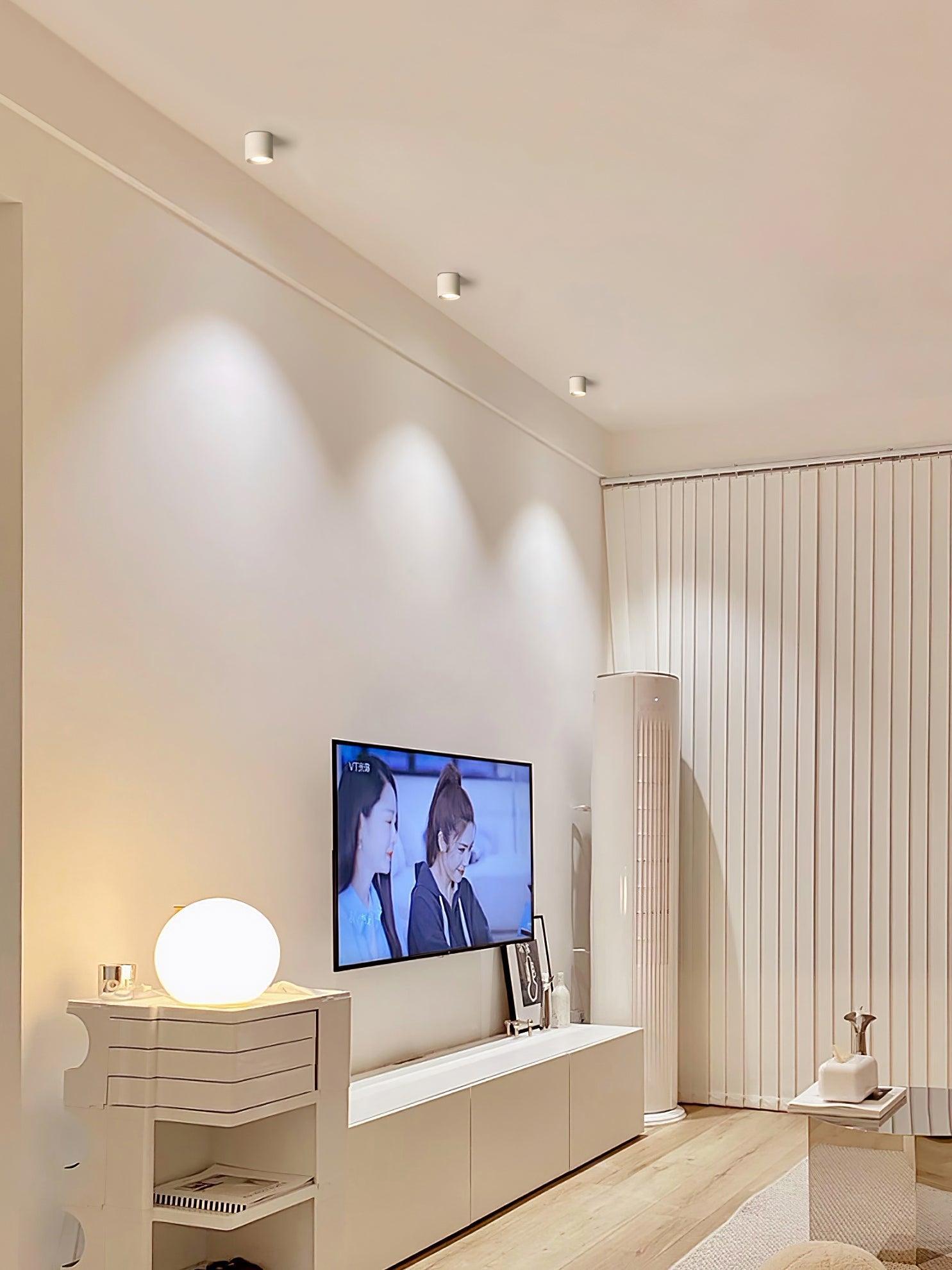 Wider TV-wall view with three white ceiling lights, round table lamp, cabinets, tall air conditioner and coffee-table edge