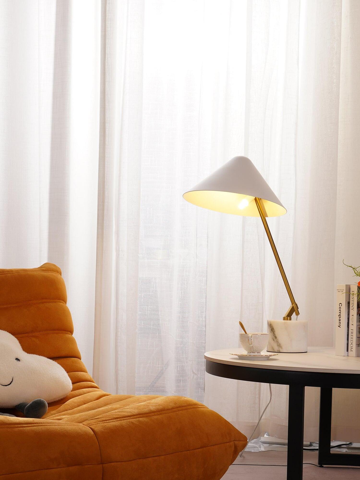 White lamp on a small round table beside an orange chair and sheer curtains