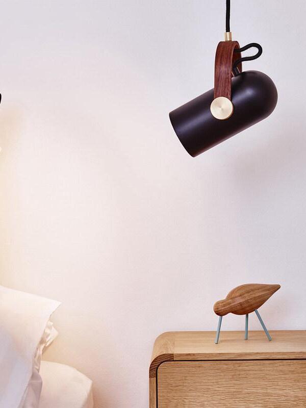 Tilted black cylindrical pendant above a wooden bedside cabinet and small bird ornament, with white pillows at left