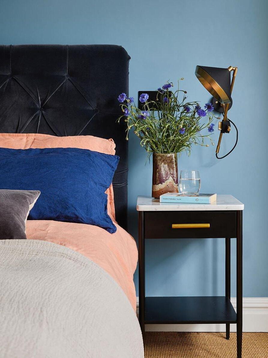 Black cone wall light beside a tufted headboard and a flower-filled bedside table on a blue wall