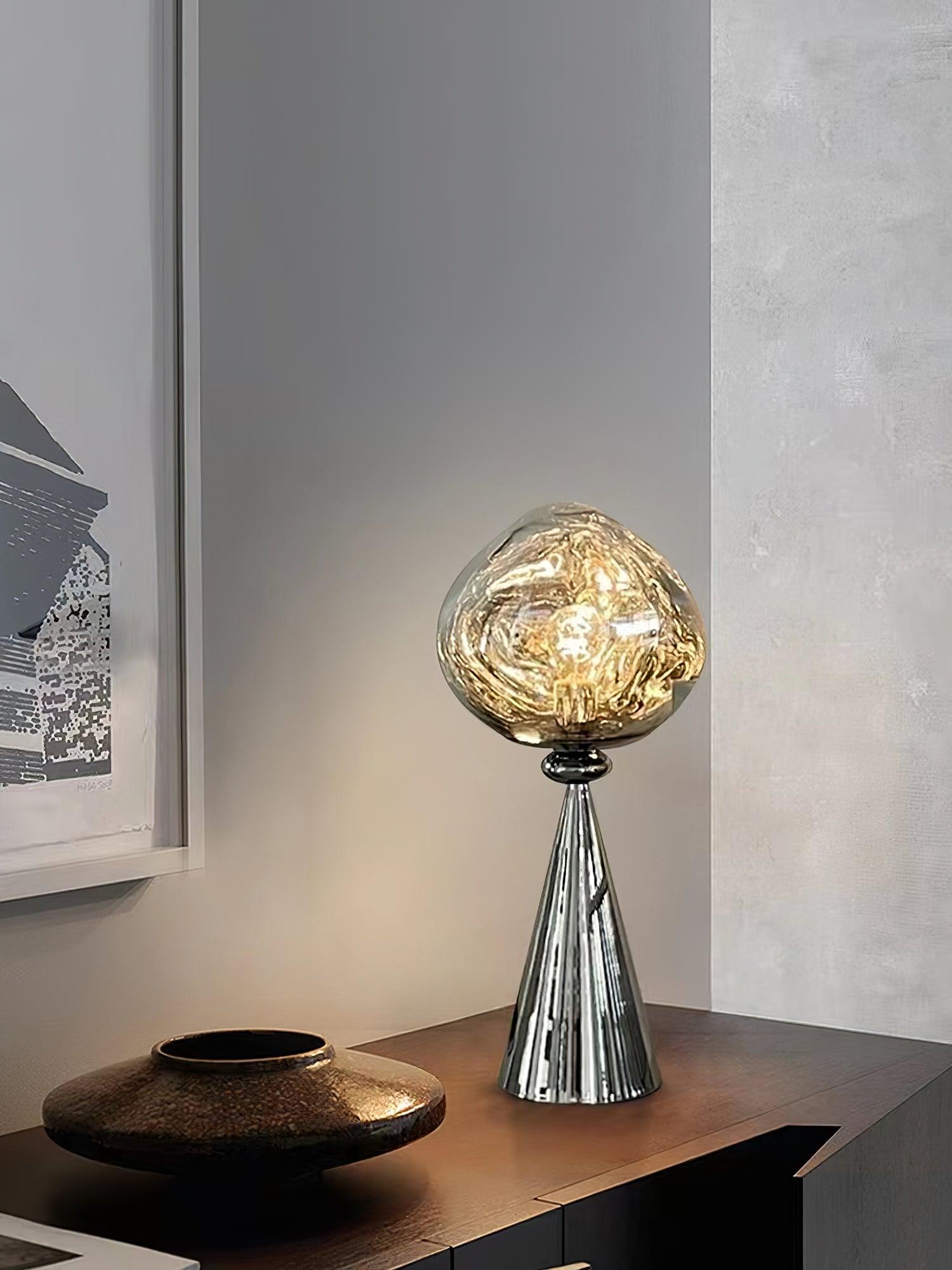 Chrome globe table lamp glowing on a wood sideboard beside a dark vessel and framed artwork
