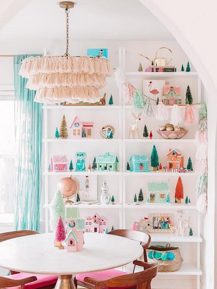 Pale blush tiered tassel chandelier above a white dining table and pink-seated chairs beside shelves of miniature houses and trees