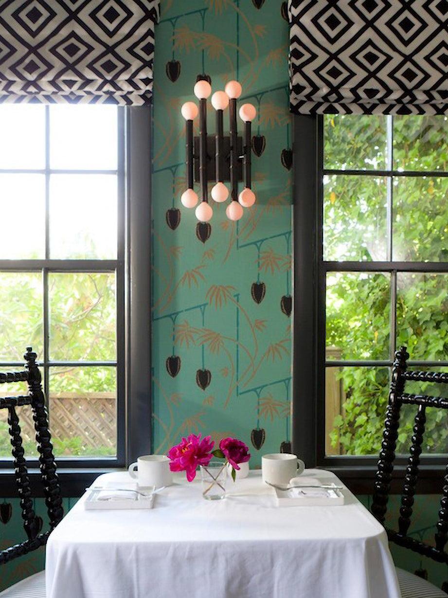 Dark Meurice wall light glowing between windows on a teal patterned wall above a small dining table and flowers
