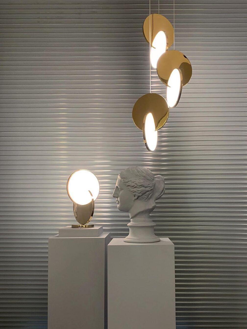 Three lit gold disc pendants and a separate table lamp beside a classical bust on plinths