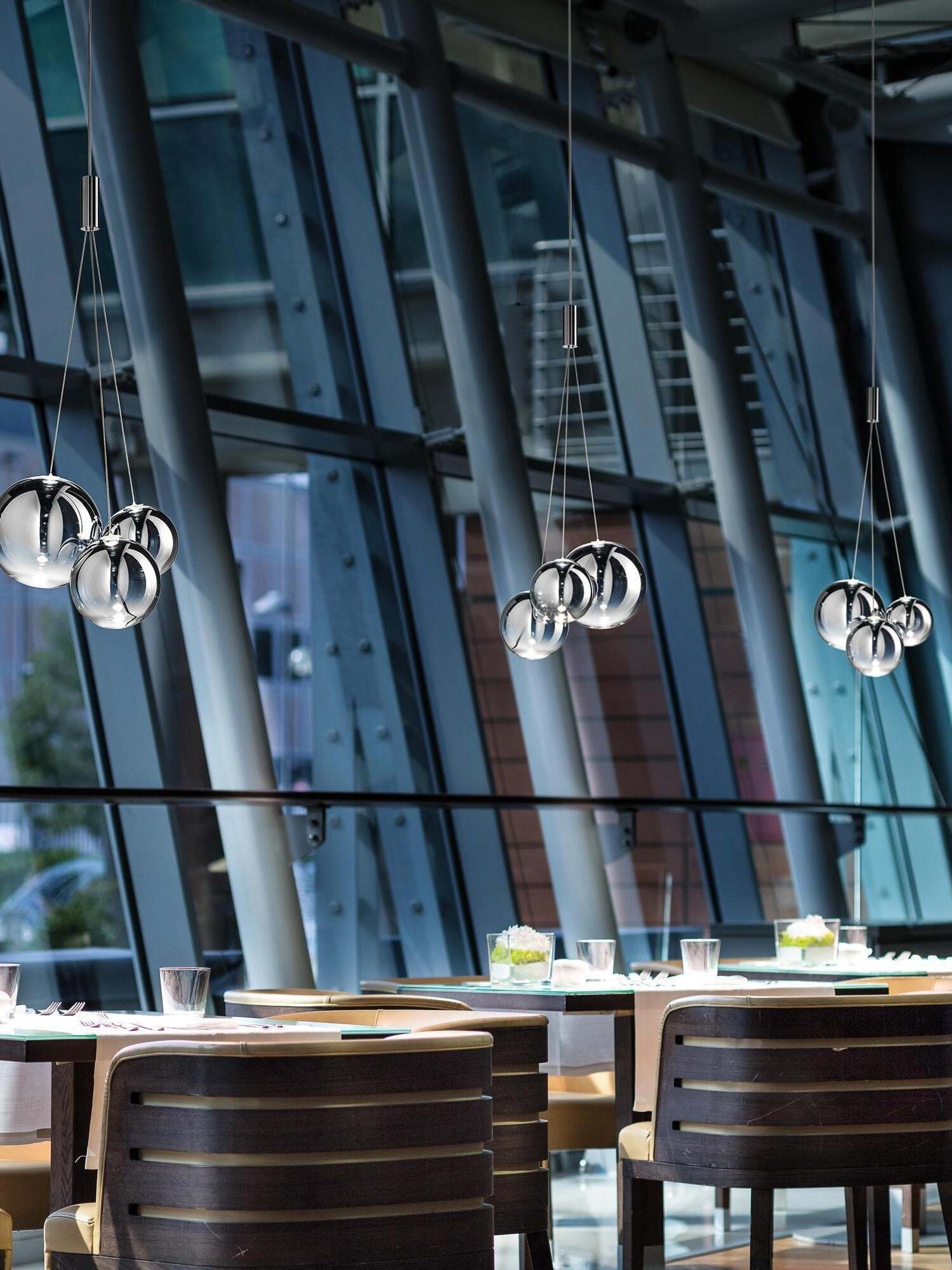 Three Smoke bubble pendants above restaurant tables with glassware, cutlery and wooden chairs beside a tall glass wall