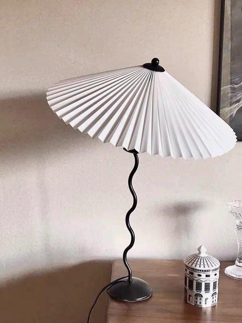 White pleated-shade Squiggle lamp on a wooden cabinet beside a ceramic miniature building