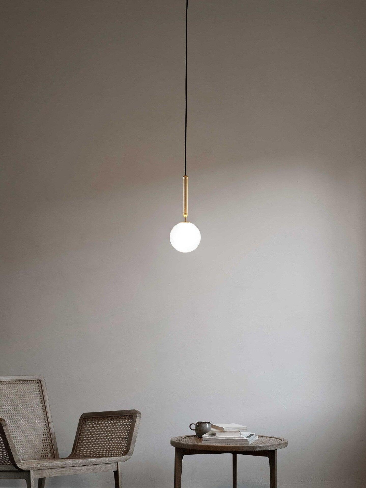 Single white globe pendant with brass-colour stem over a woven chair and round coffee table with books
