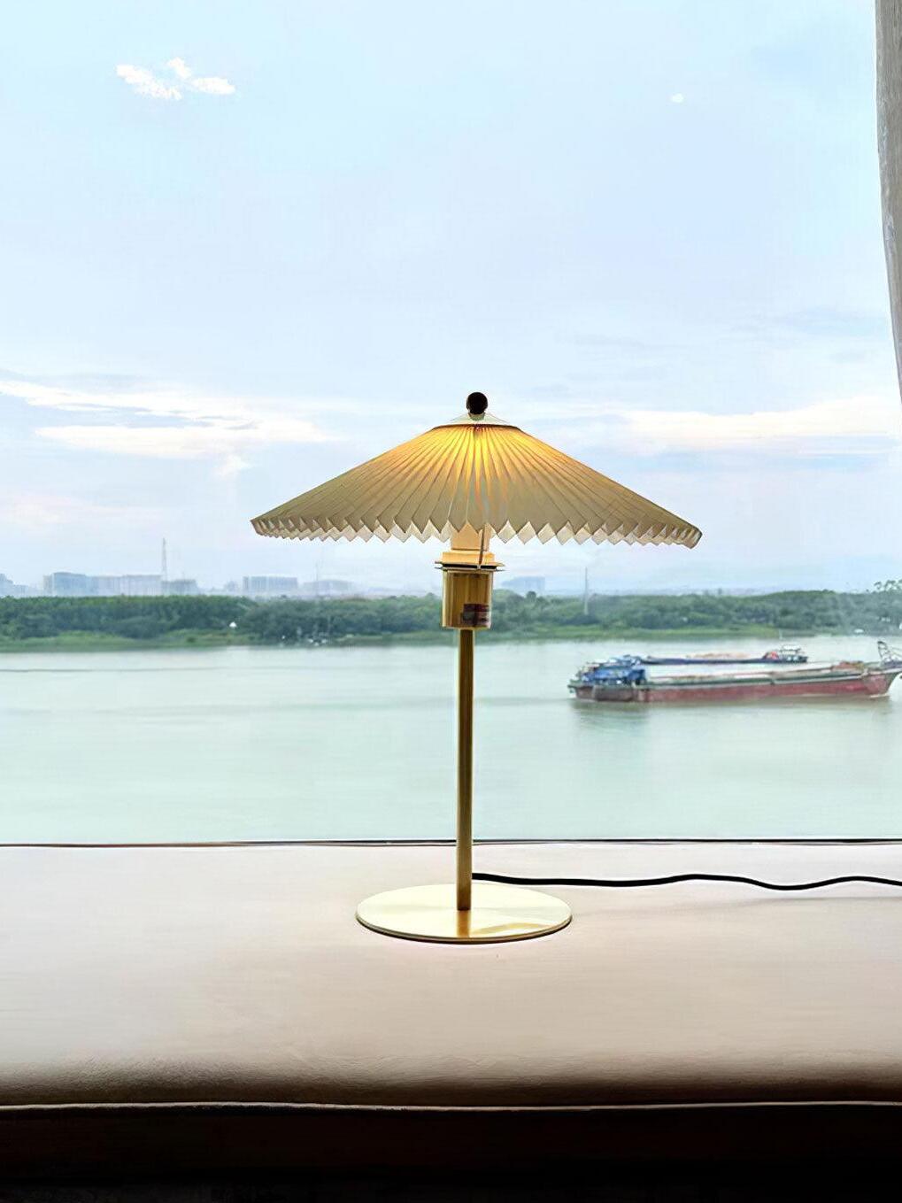 Pleated table lamp on an indoor window ledge overlooking water, with its cable visible to the right