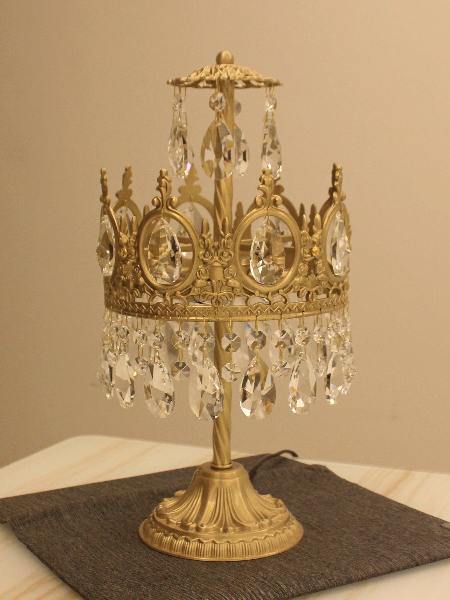 Unlit crystal table lamp on a dark mat, showing the upper drop arrangement and gold-tone round base.
